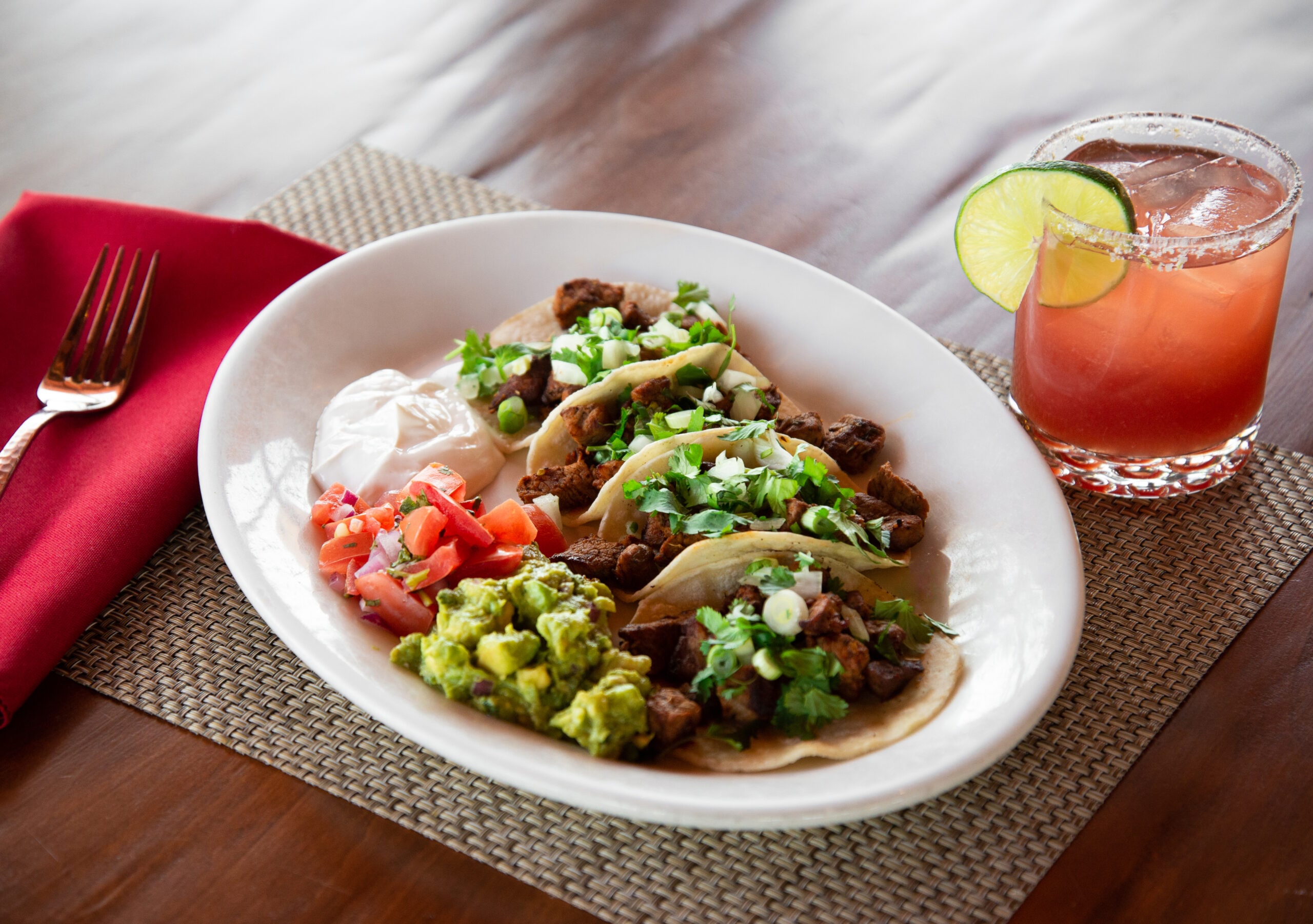 Photo of four beef tacos on a white plate served with cilantro, onions, pico de gallo, guacamole, and sour cream next to a red cocktail with a lime and salted rim.