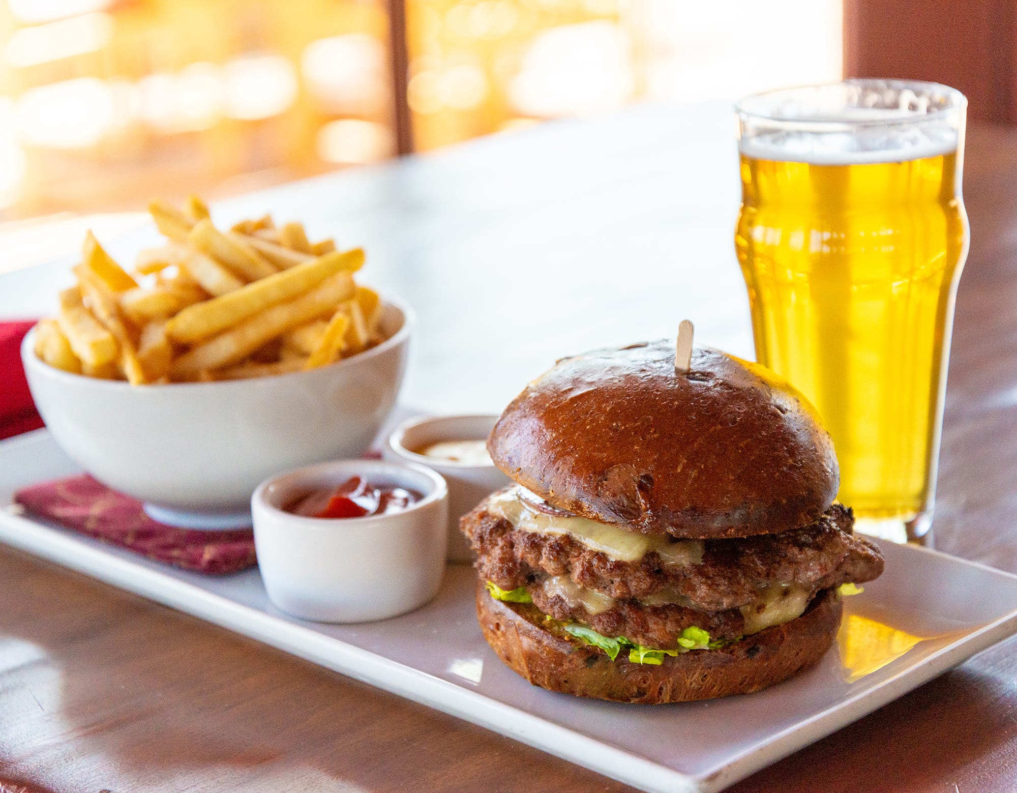 A photo of a cheese burger, fries, and ketchup sitting on a long, white platter and a cup of golden beer on a brown table.