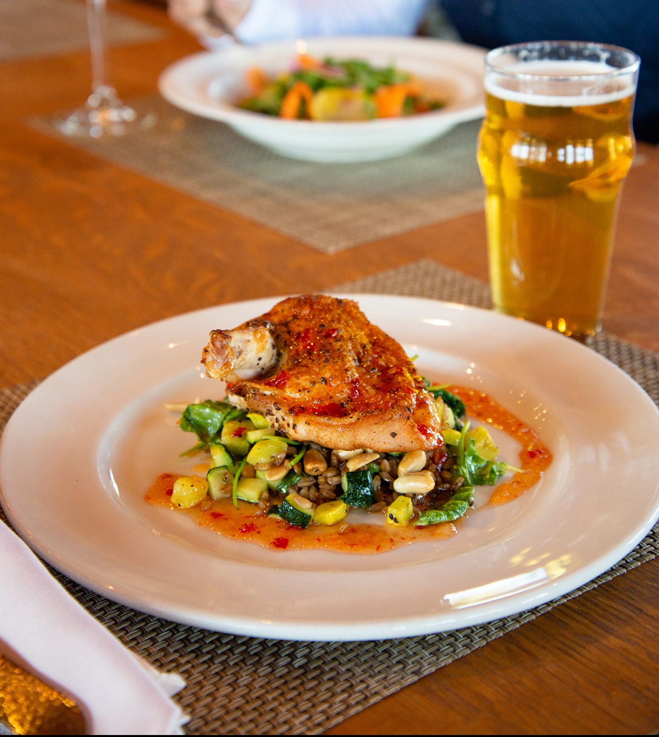 Photo of a plate filled with seasoned chicken on top of a green and seseme seed salad next to a cup of golden beer on a brown table.