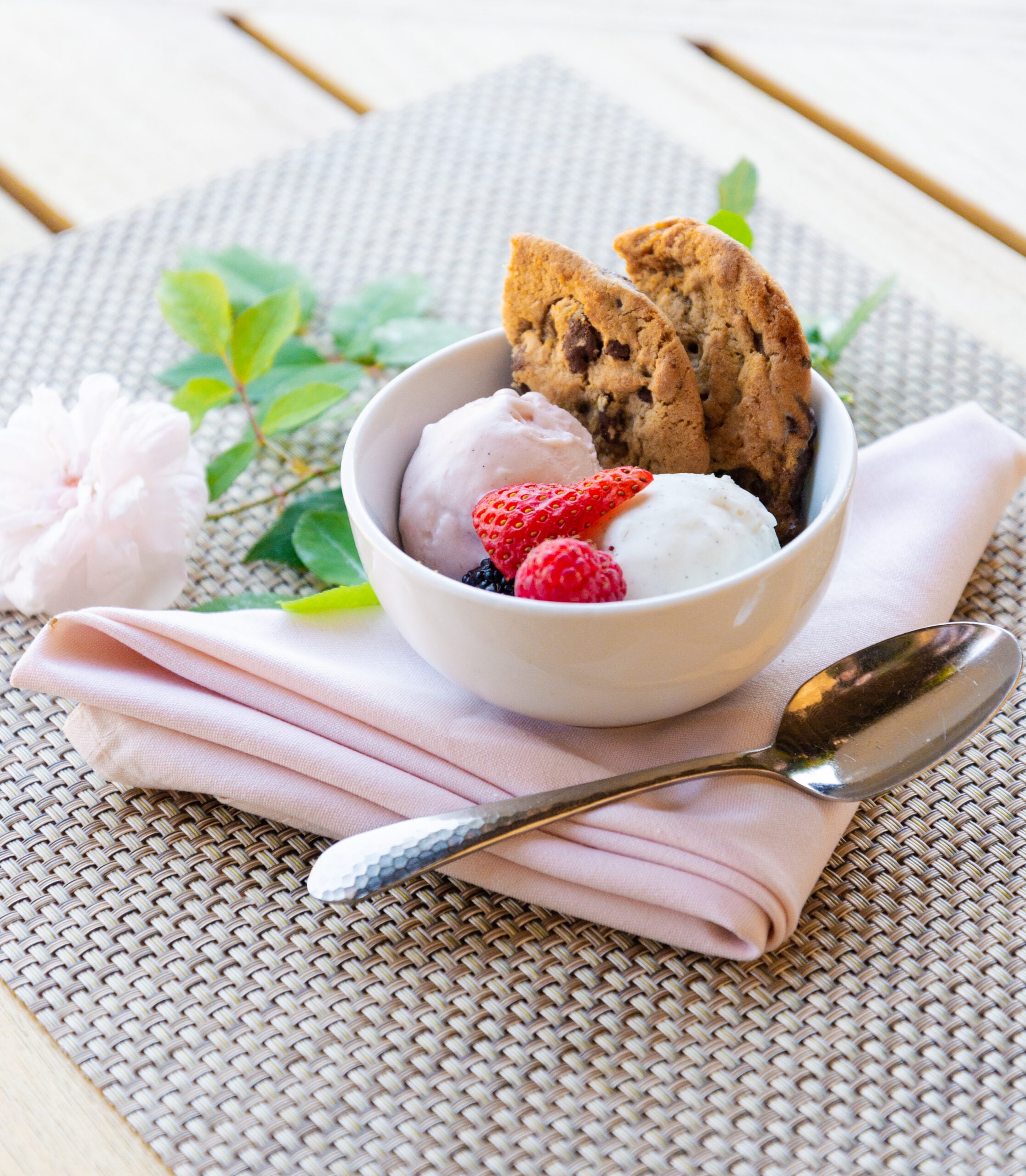 Photo with a bowl of ice cream with two chocolate chip cookies and raspberries sitting on top of a pink napkin next to a green leaves, pink flower, and silver spoon.