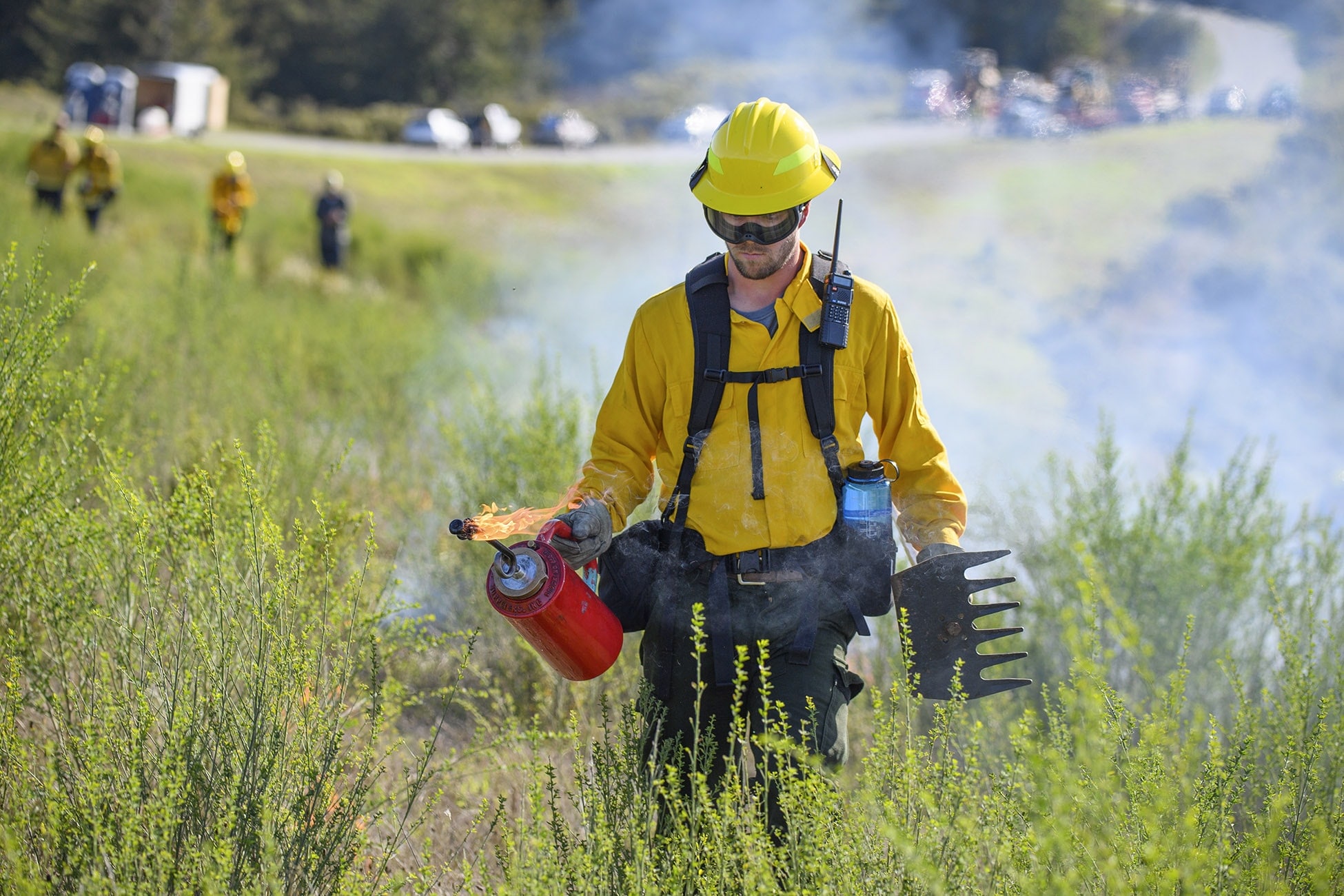 A person wearing yellow protective equipment while holding a weed removal tool and a controlled flame coming from a red chamber walking through green brush.