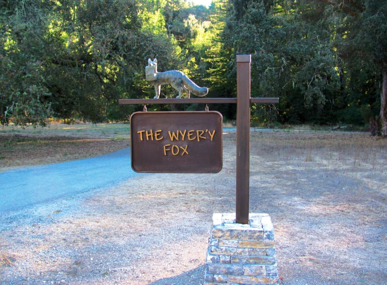 A front yard sign that has a sculpture of a fox holding a bag on top of a sign that has The Wyer'y Fox.